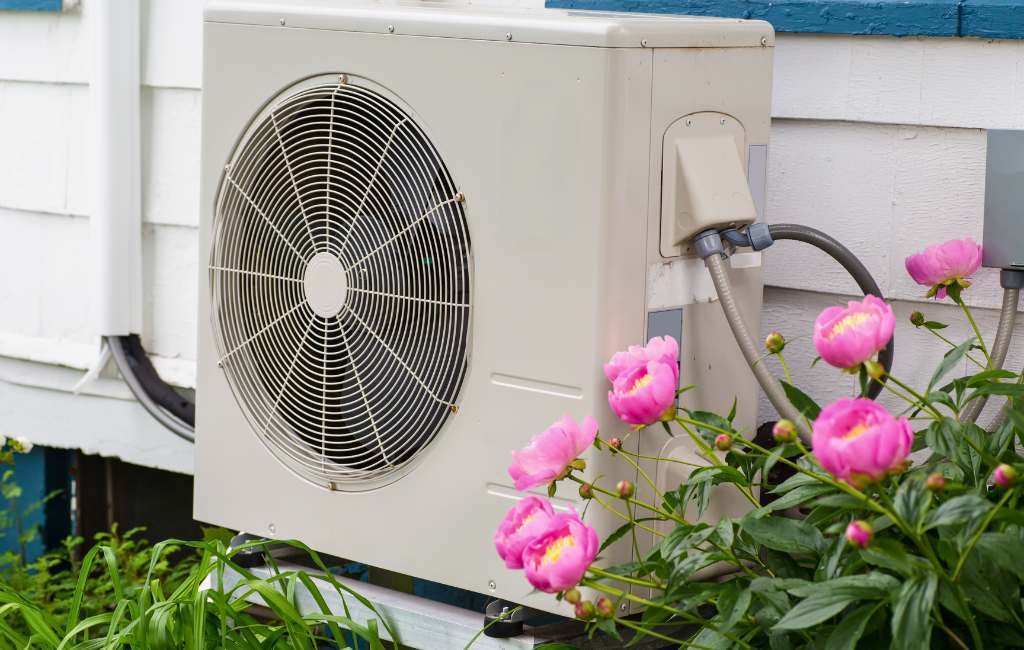 Wärmepumpen-Außengerät neben blühenden Blumen vor einem Wohnhaus