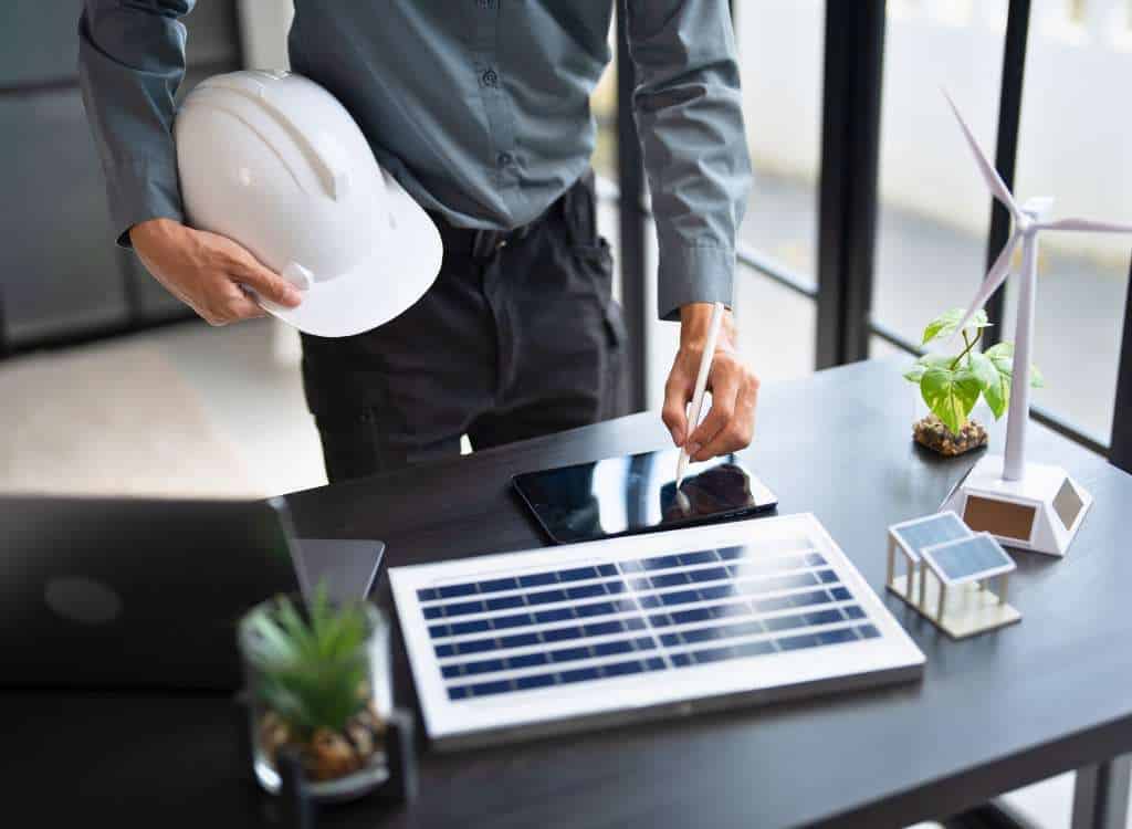 Techniker mit Helm und Tablet analysiert Photovoltaikmodul und Energiedaten auf einem Schreibtisch.
